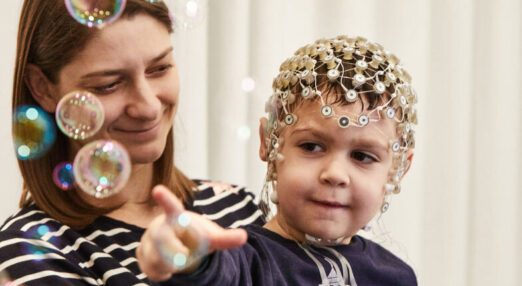 A mother holds a child who points at bubbles, while being monitored using a full head brain imaging device.