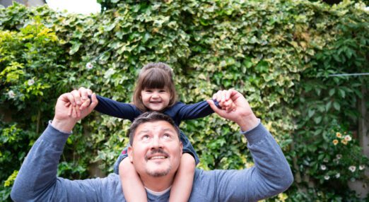 A man holds a young girl on his shoulders and looks up towards her