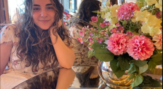 Sophie sits smiling at a table. There is a with a vase of flowers.