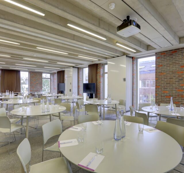 Round tables with chairs around each, filling a room which has large windows spaced around the side