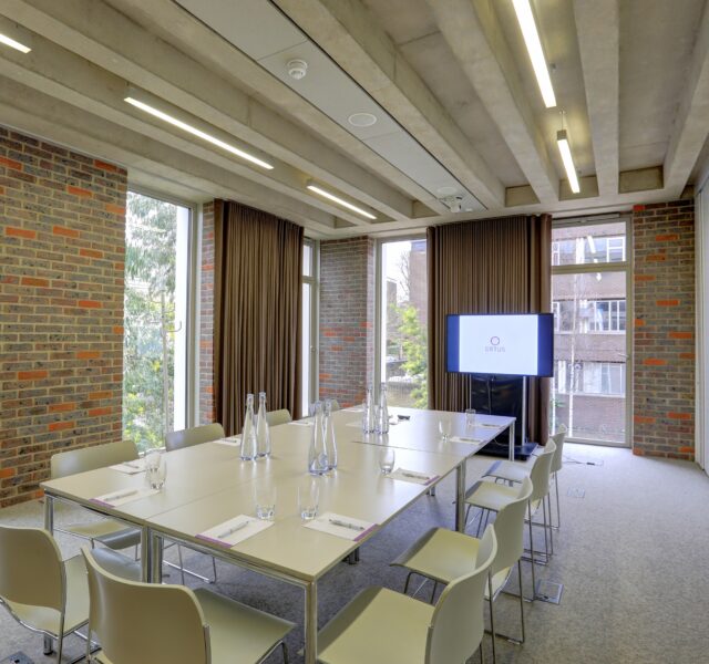 A boardroom layout of tables and chairs, with 10 people seated around a rectangular table layout, all within a room with tall windows