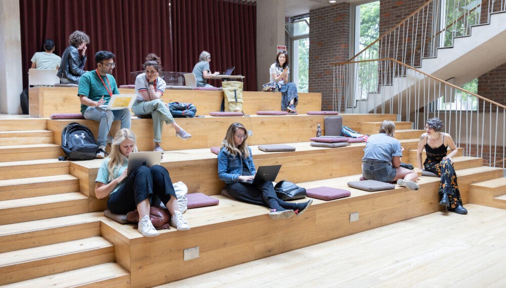 A room space with four levels of wood, with cushions laid out. A few people are seated on the cushions, working on laptops or having a conversation