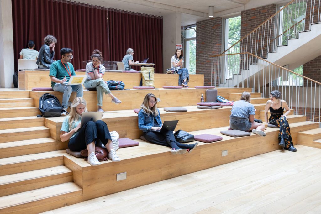 A room space with four levels of wood, with cushions laid out. A few people are seated on the cushions, working on laptops or having a conversation