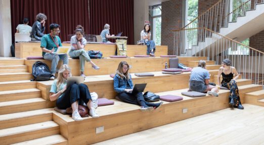 A room space with four levels of wood, with cushions laid out. A few people are seated on the cushions, working on laptops or having a conversation