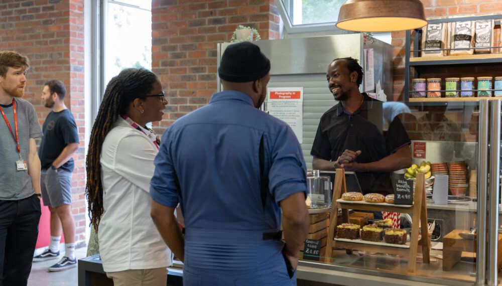 A man standing behind a cafe counter, with cakes visible on plates. Two people are seen from behind, ordering and talking with the cafe staff member