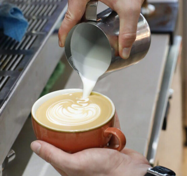 Milk being poured from a metal jug in to a coffee , creating a flower-shaped pattern