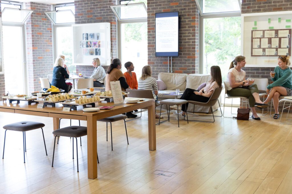 Trays of sandwiches and finger food waiting on a table, with small groups of people seated, talking together behind