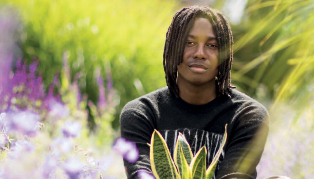 A young black man sitting among plants, holding a pot plant in both hands in front of him