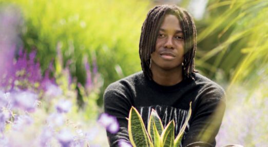 A young black man sitting among plants, holding a pot plant in both hands in front of him