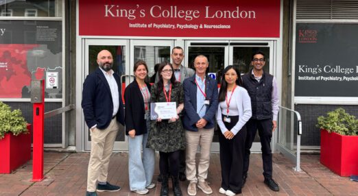 A small group of 7 people, standing outside the doors of King's College London, holding a certificate proudly