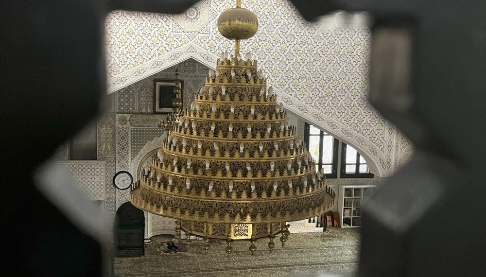 An interior of a detailed and ornate mosque, showing a large chandelier and pattered walls and floor. This is seen through a hole, shaped as an eight pointed star