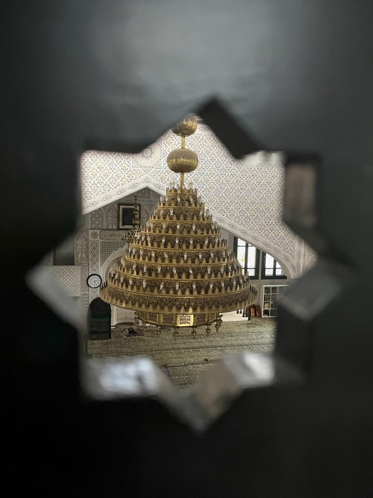 An interior of a detailed and ornate mosque, showing a large chandelier and pattered walls and floor. This is seen through a hole, shaped as an eight pointed star