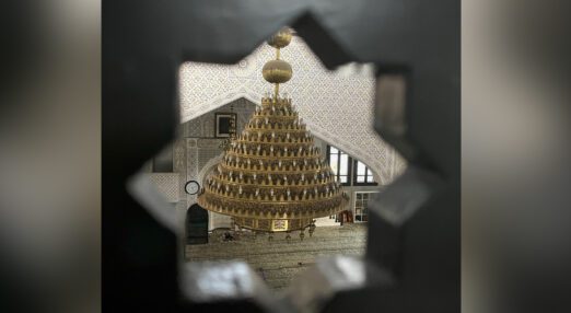 An interior of a detailed and ornate mosque, showing a large chandelier and pattered walls and floor. This is seen through a hole, shaped as an eight pointed star