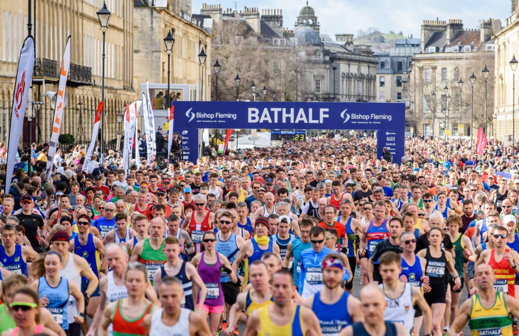 Many runners together, passing between beige stone buildings. In the background is a sign stating Bath Half