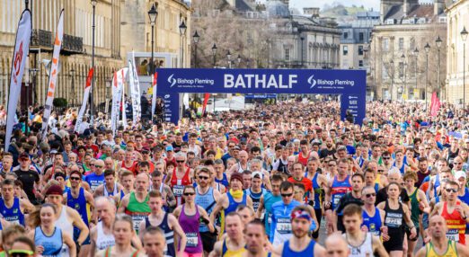 Many runners together, passing between beige stone buildings. In the background is a sign stating Bath Half