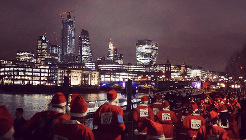 Many people dressed in Santa costumes, moving beside the Thames at night, with a London skyline in the backgroun