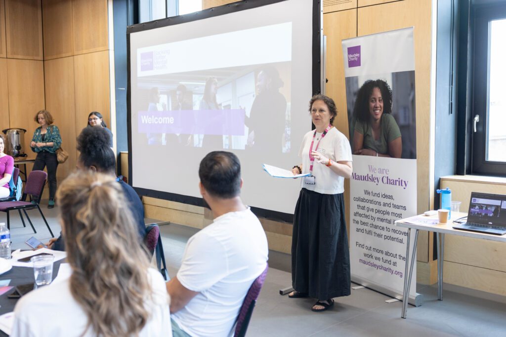 A woman (Georgina Chatfield) standing beside a large screen, with the words Welcome visible. Attendants of an event are visible to the side and the woman is speaking to the room.