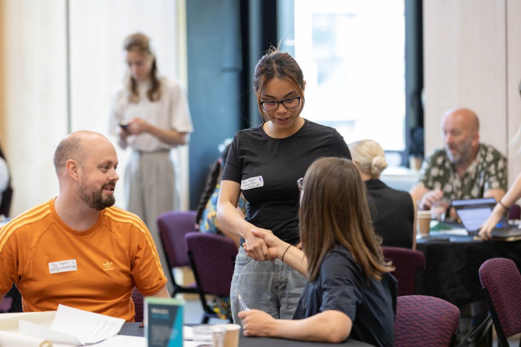 A standing young woman shaking hands with a woman who is seated. A man is sitting beside them. Two people have visible name badges explaining who they are which organisation they work for.
