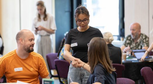 A standing young woman shaking hands with a woman who is seated. A man is sitting beside them. Two people have visible name badges explaining who they are which organisation they work for.