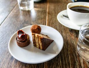 A small plate with three small different pieces of cake on it, sitting on a wooden table top, beside a cup of coffee in a cup and saucer