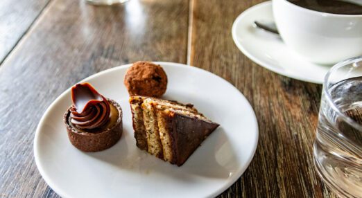 A small plate with three small different pieces of cake on it, sitting on a wooden table top, beside a cup of coffee in a cup and saucer