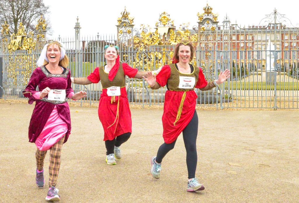Three women dressed up in Tudor clothing, running in an event in front of Hampton Court Palace, the gates and building visible