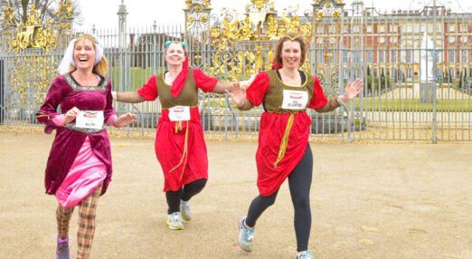Three women dressed up in Tudor clothing, running in an event in front of Hampton Court Palace, the gates and building visible