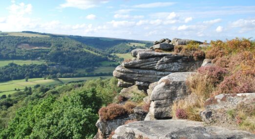 The view from on top of a hill with large rocky outcrops, looking down over a green valley of trees and fields