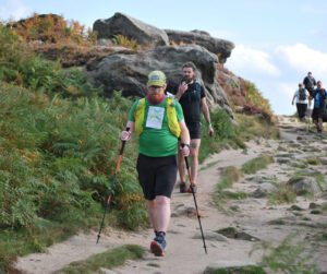 A man walking along a dirt path using walking poles. An even bib with competitor number is on his front. There is a man walking close behind him and the path passes large rocks and some plants