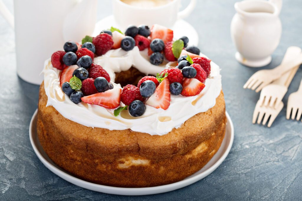 Cream and fruit covered sponge cake, sitting on a plate, on a table with forks