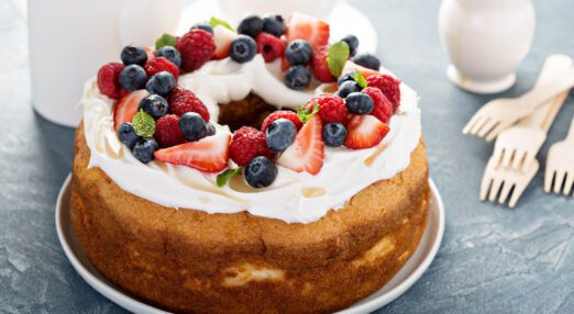 Cream and fruit covered sponge cake, sitting on a plate, on a table with forks