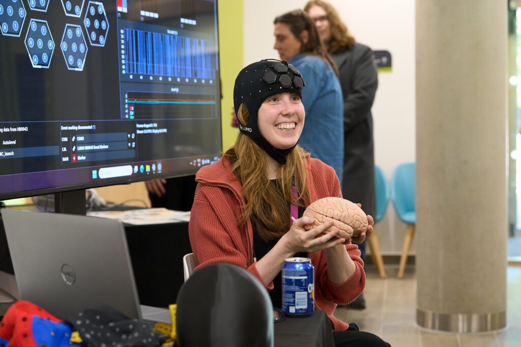 A young woman seated indoors, holding a brain replica, wearing a head dress which appears to have sensors in-built. The woman is smiling excitedly. Behind her is a large screen with data and analysis visible.