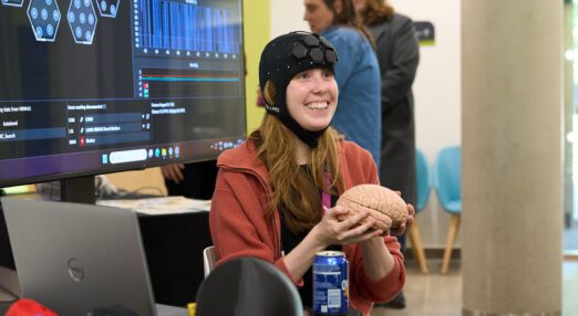 A young woman seated indoors, holding a brain replica, wearing a head dress which appears to have sensors in-built. The woman is smiling excitedly. Behind her is a large screen with data and analysis visible.